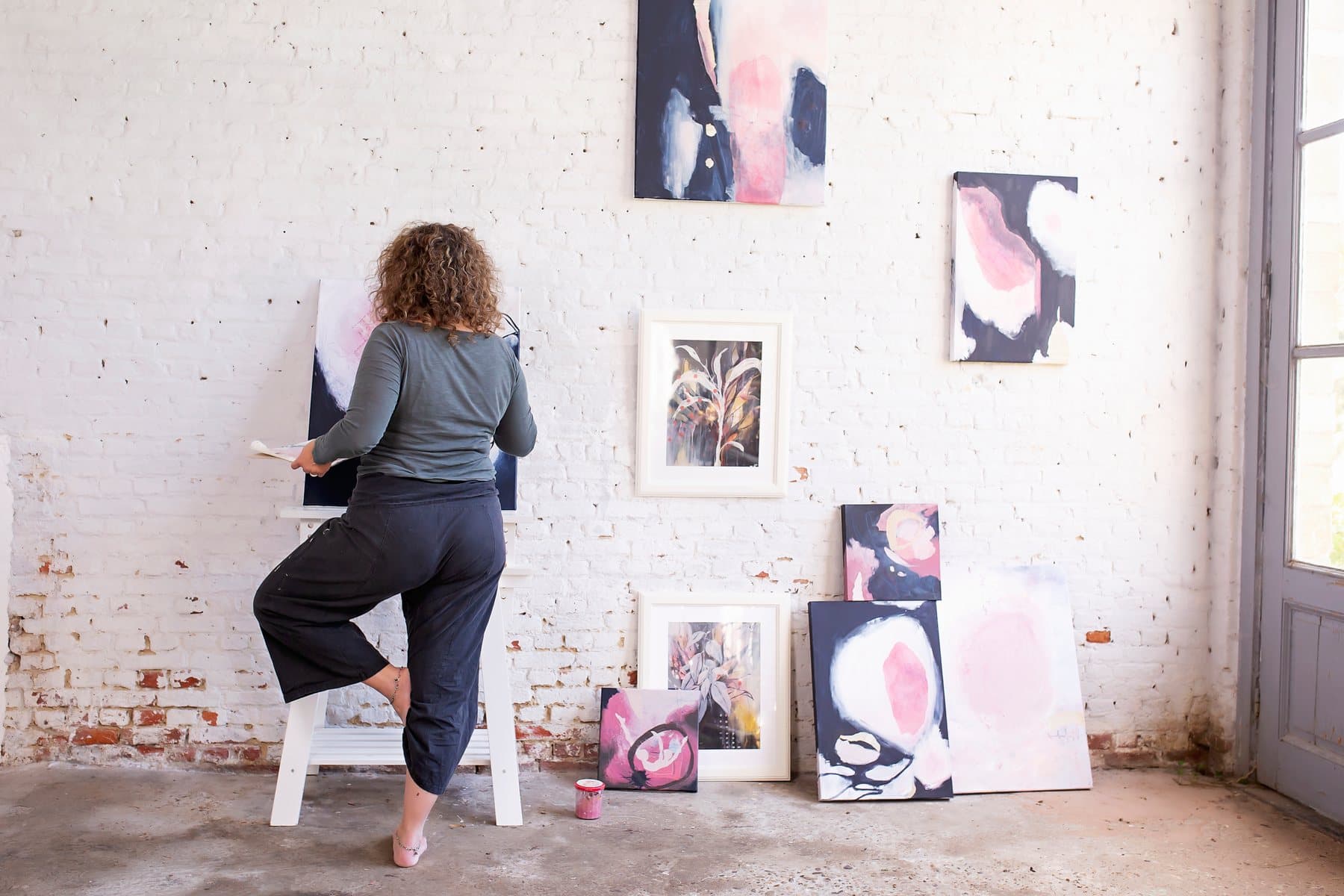 Mieke Fleurackers standing at her easel in front of a white brick wall hung with her paintings